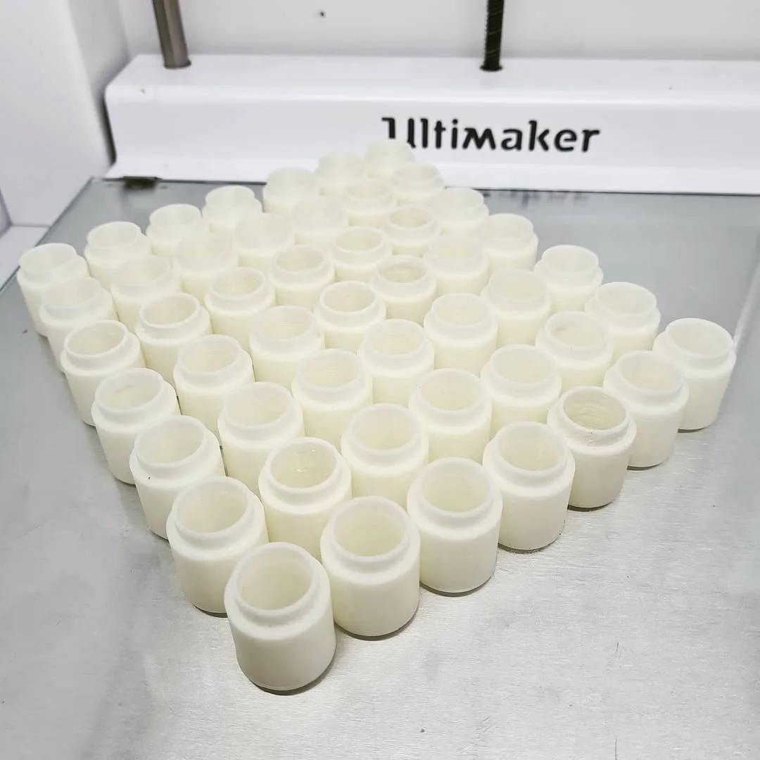 Mini serija istovetnih belih 3D štampanih rezervnih delova od ABS-a, poređanih ispred štampača Ultimaker